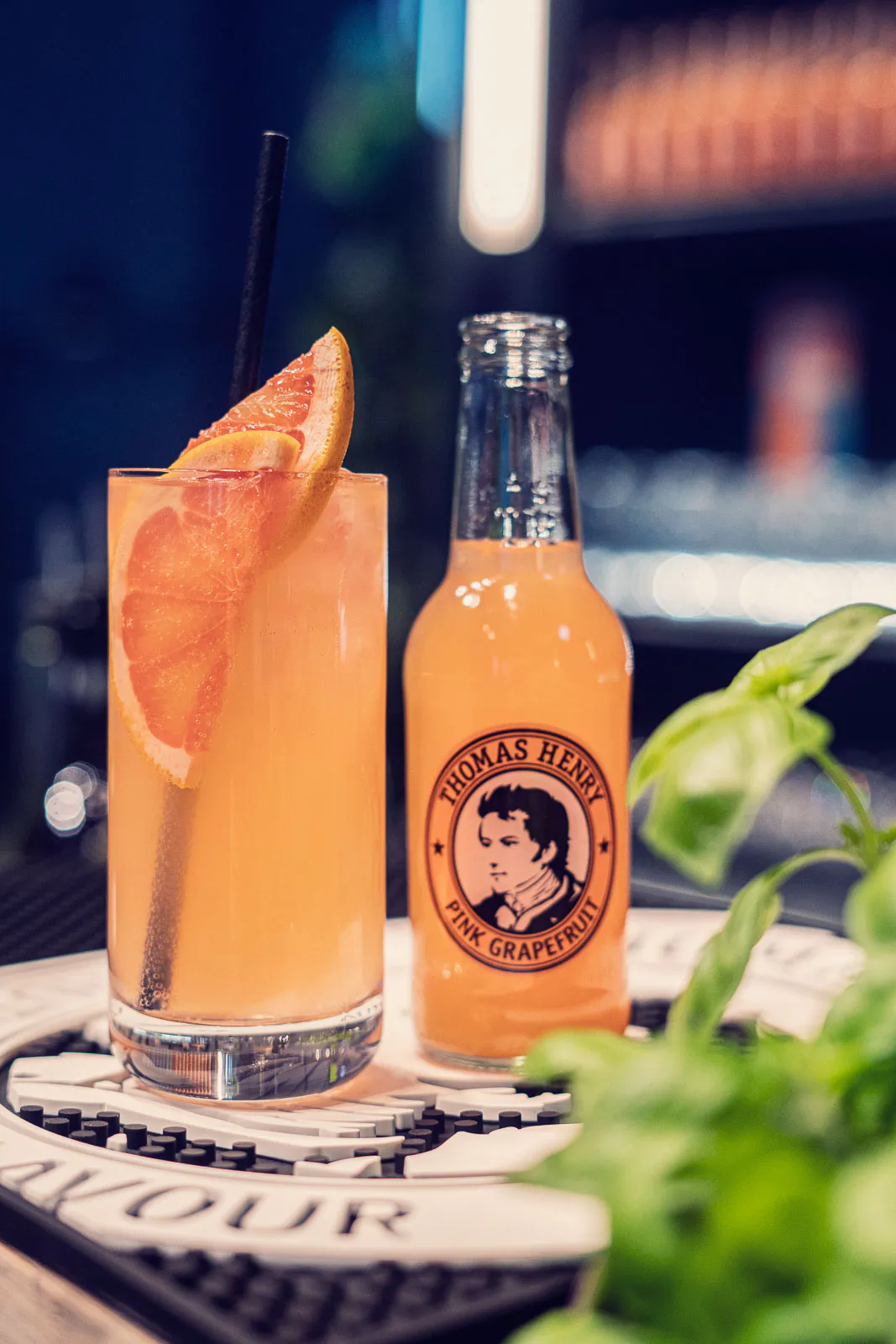 Orangefarbener Drink mit Grapefruit und Strohhalm neben einer Thomas Henry Pink Grapefruit-Flasche auf einer Bartheke.