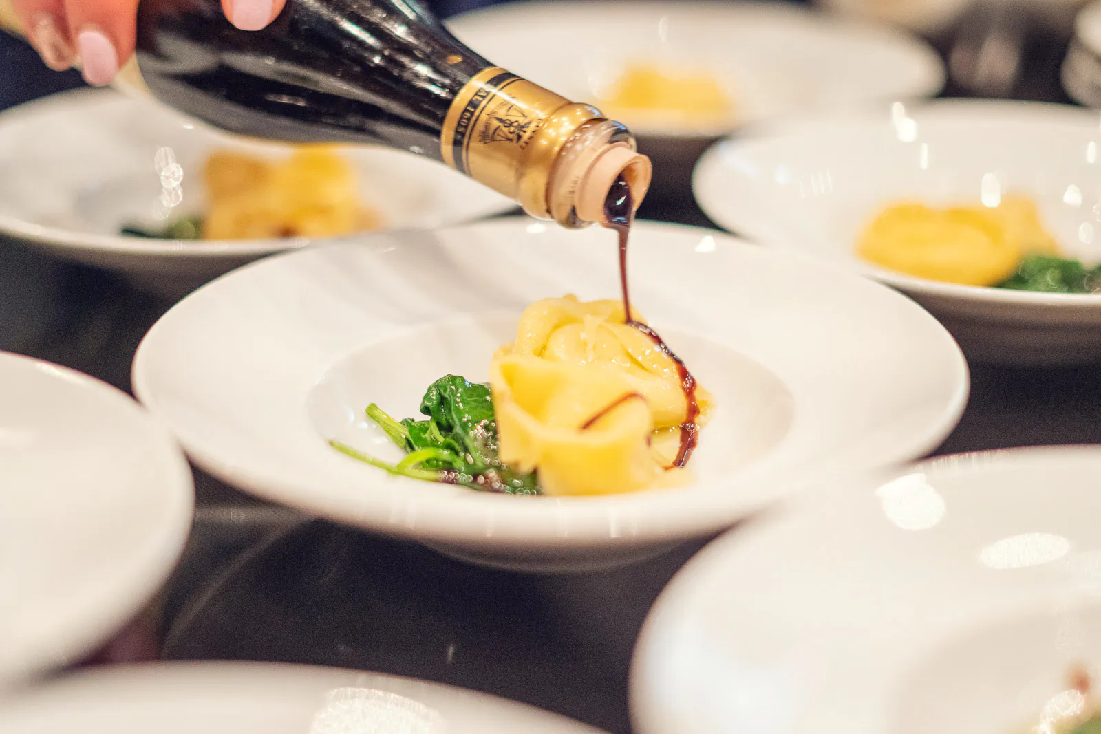 Eine Hand gießt dunkle Sauce aus einer Flasche über gefüllte Pasta mit grünem Gemüse auf einem weißen Teller.