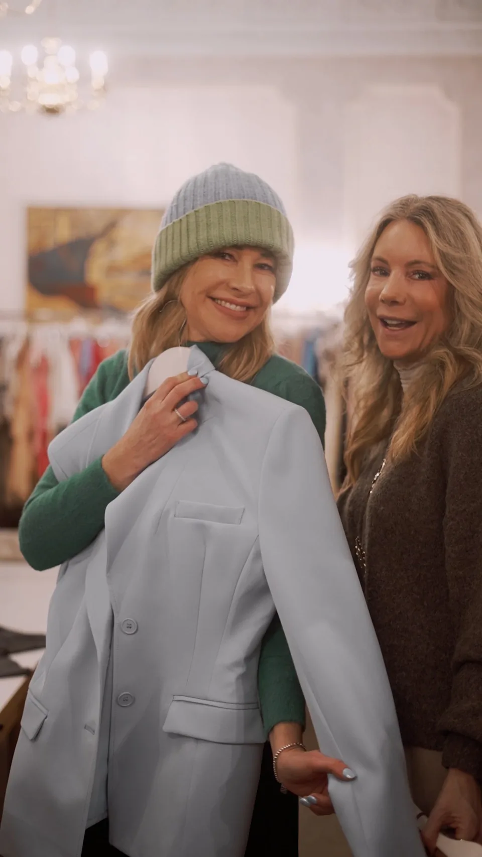 Zwei lächelnde Frauen in einem hellen Showroom, eine hält einen hellblauen Blazer vor sich und trägt eine Strickmütze.