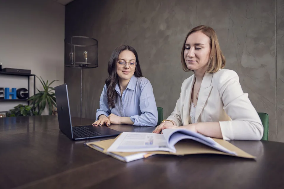 Zwei Frauen sitzen im Büro an einem dunklen Tisch mit Laptop und aufgeschlagener Mappe vor einer grauen Wand.