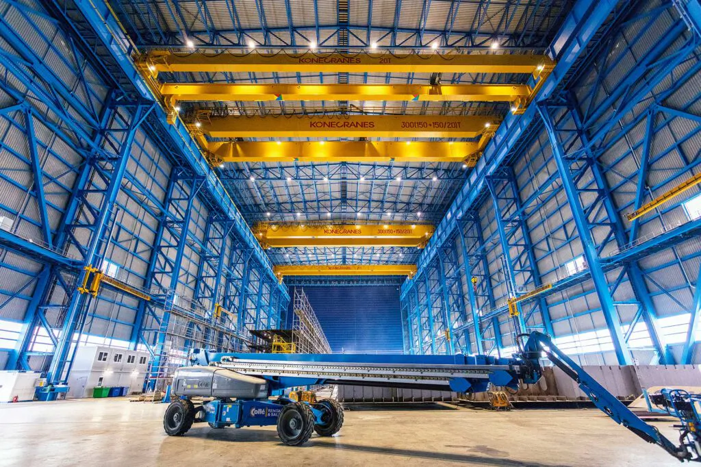 Gelbe Konecranes-Brückenkräne unter blauem Stahltragwerk einer lichtdurchfluteten Industriehalle mit Arbeitsbühne.