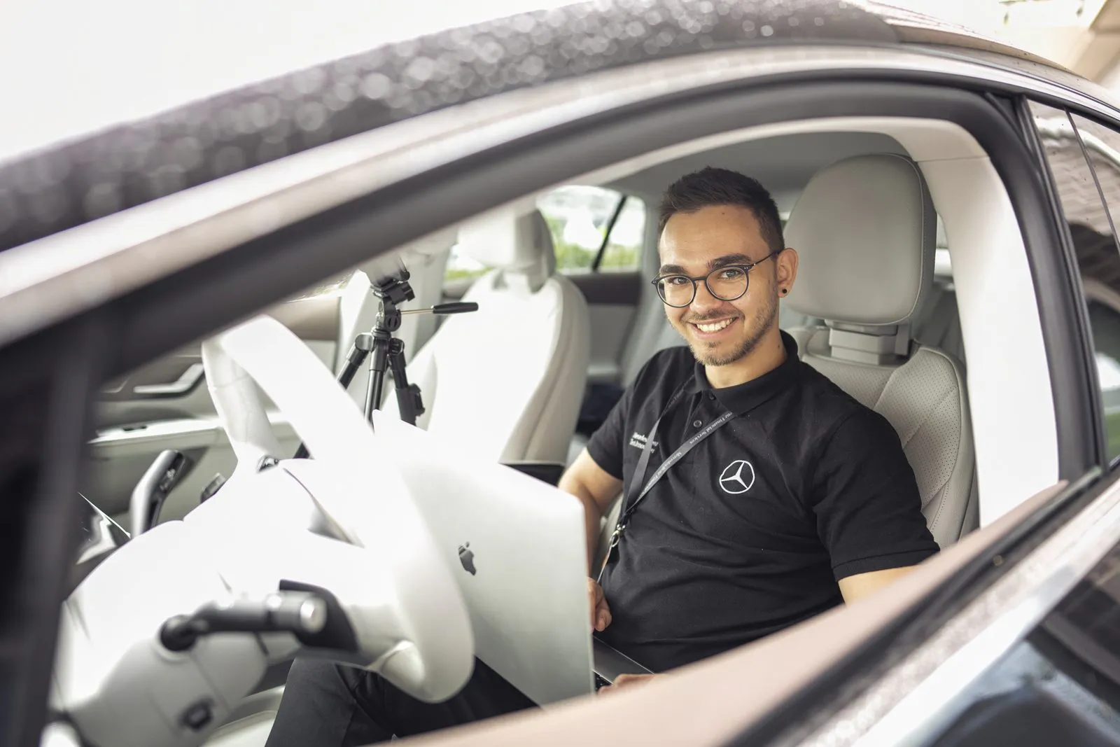 Lächelnder Mann im Mercedes-Benz-Polo sitzt mit Laptop am Steuer eines Autos mit hellem Lederinterieur.
