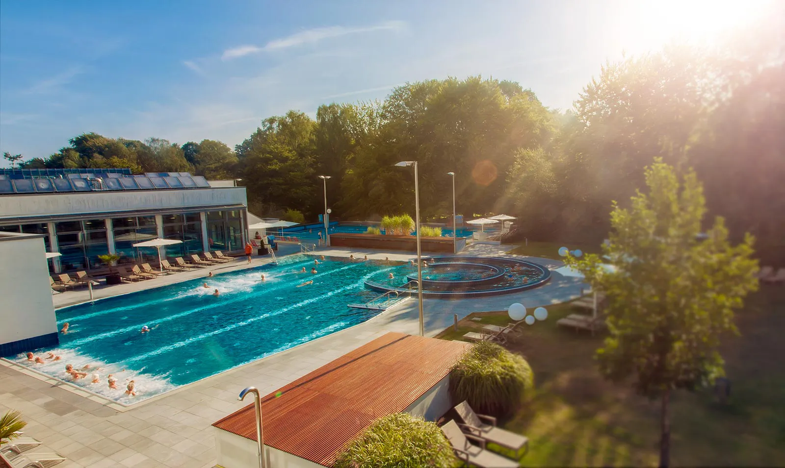 Sonniger Außenbereich von Vitasol mit Schwimmenden im türkisblauen Becken, rundem Wasserbecken und Liegestühlen.
