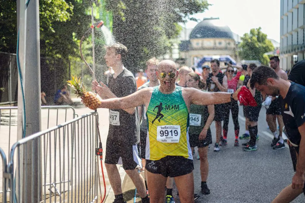 Läufer mit Ananas und ausgebreiteten Armen kühlt sich im Sprühnebel auf sonniger, abgesperrter Straße ab.