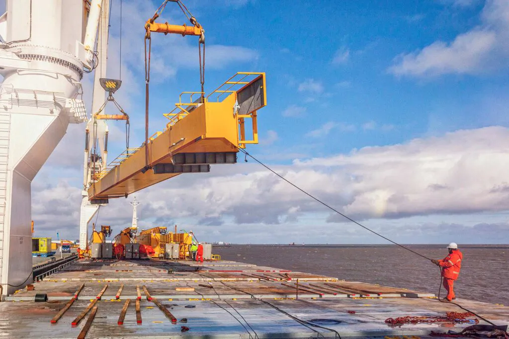 Arbeiter in orangefarbener Schutzkleidung führt einen schwebenden gelben Kranträger per Seil auf einem Schiffsdeck.