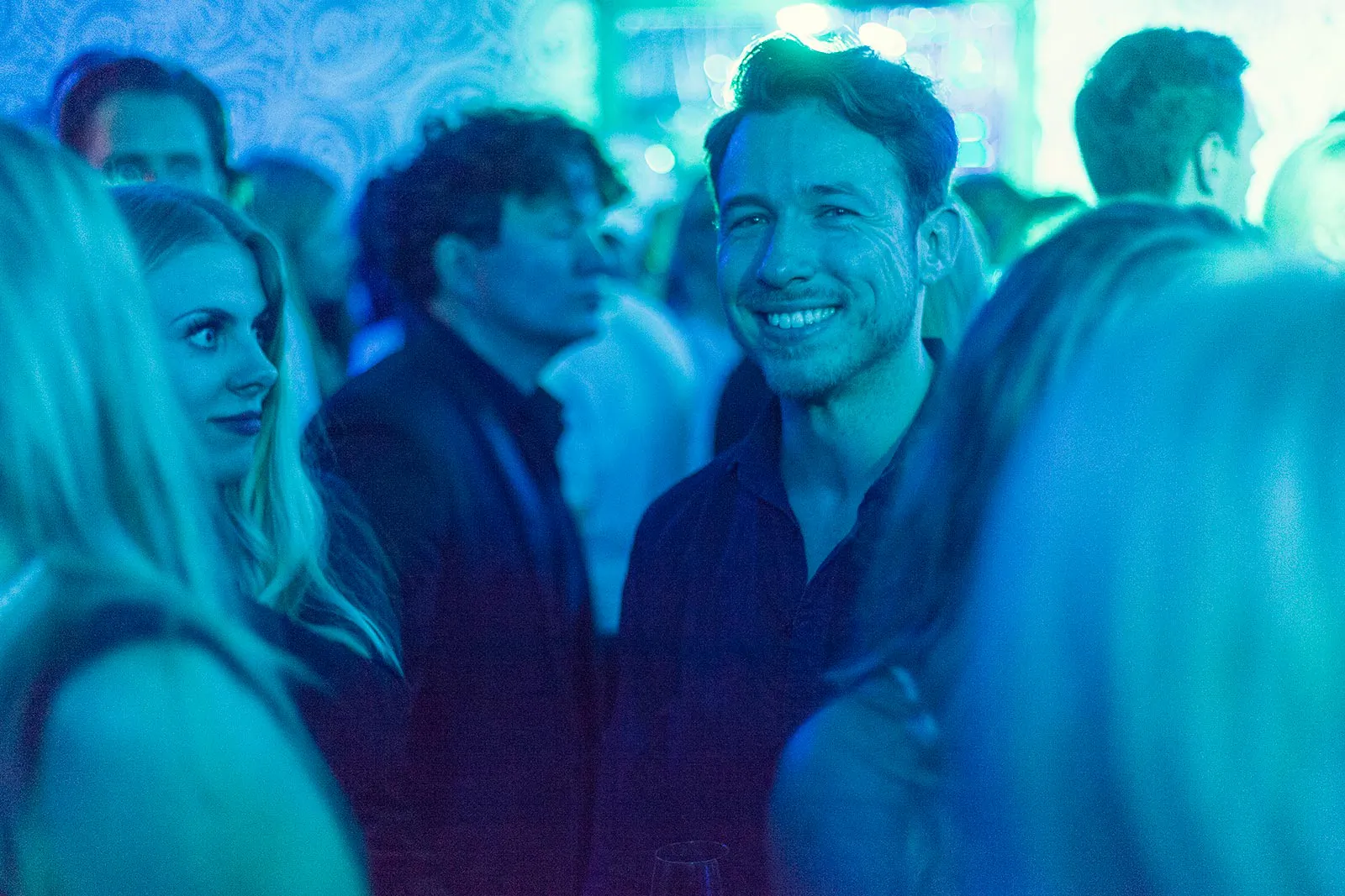 Lächelnder Mann mit Sonnenbrille auf dem Kopf zwischen Partygästen in blau-türkisem Licht im Innenraum.