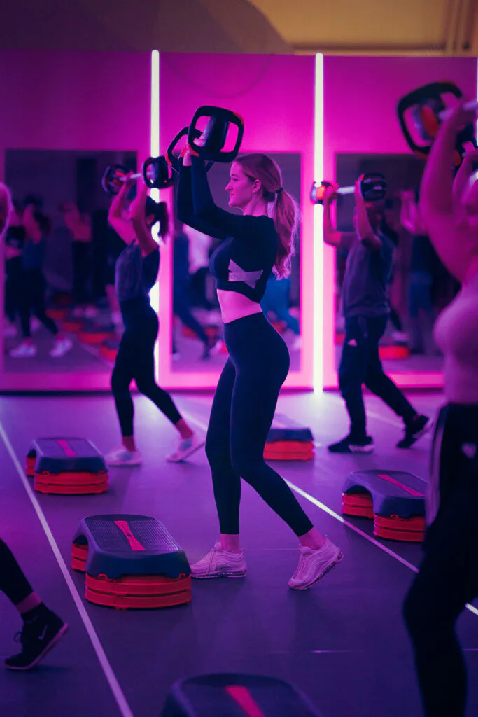 Teilnehmende heben Gewichte im Fitnesskurs der Energy Arena, umgeben von Steppern und pinkem Licht.