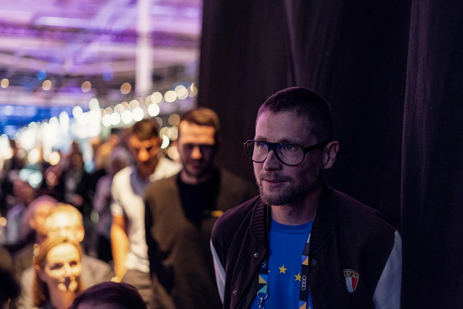 Mann mit Brille vor schwarzem Vorhang, dahinter Gäste in einer violett beleuchteten Eventhalle beim MAX Award.