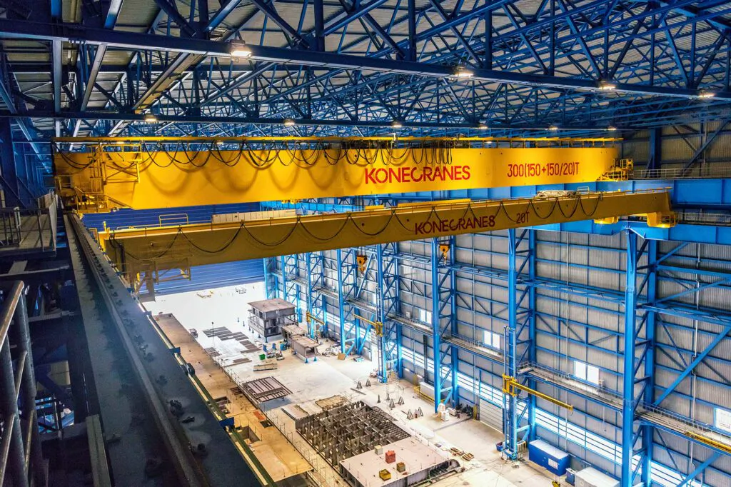 Gelbe Konecranes-Brückenkräne unter blauem Stahltragwerk in einer Industriehalle mit Tageslicht.