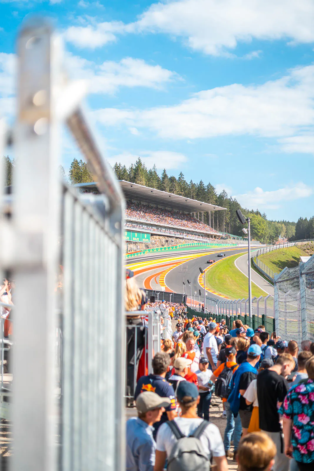 Zuschauer an einer umzäunten Formel-1-Rennstrecke mit steiler Kurve und voller Tribüne unter blauem Himmel.