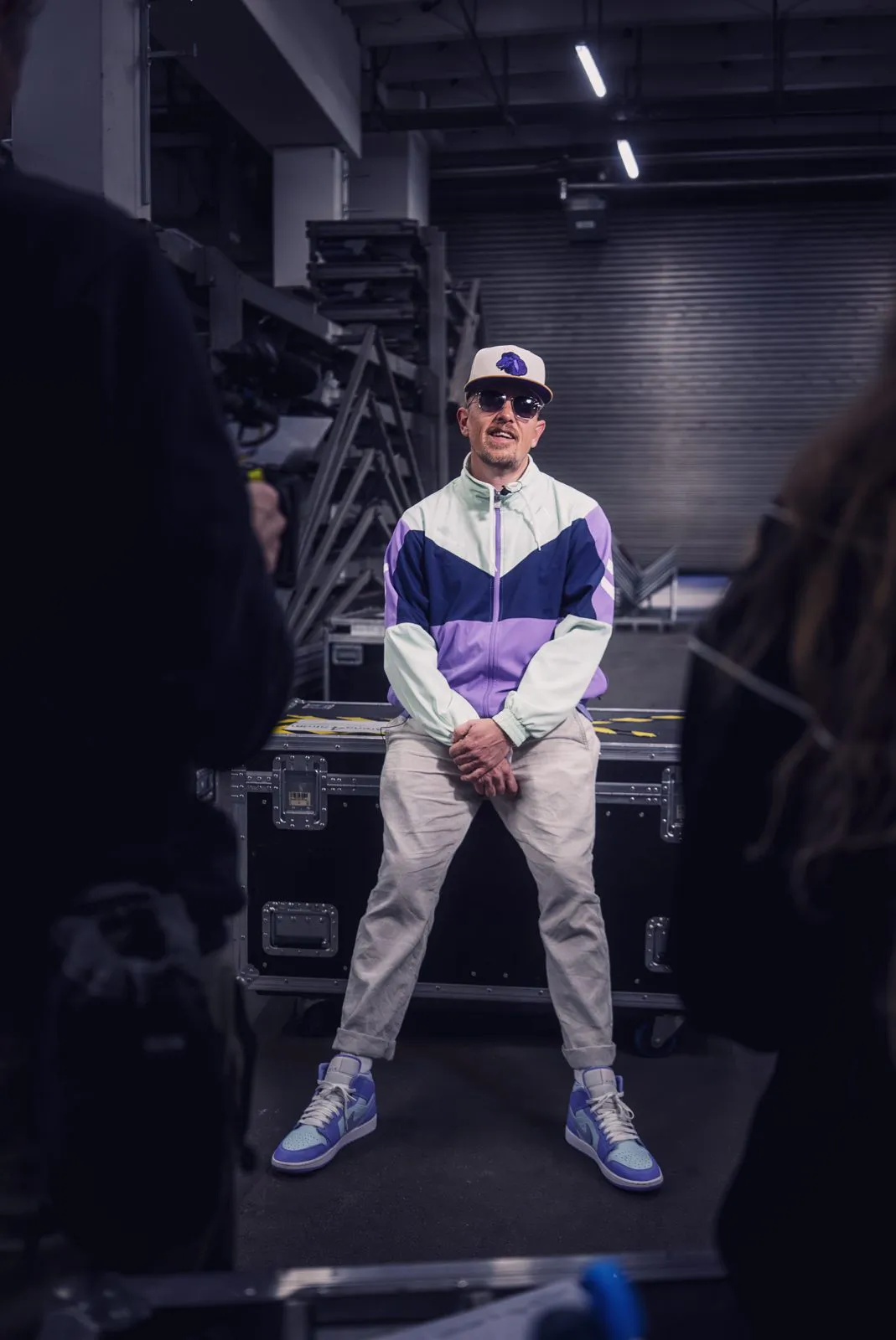 Jan Delay mit Cap, Sonnenbrille und lila-weißer Trainingsjacke vor Transportkisten im Backstage-Bereich.