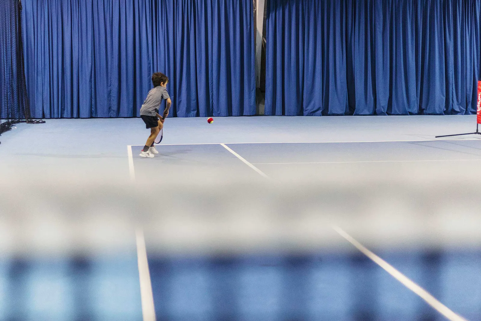 Kind mit Tennisschläger läuft beim Wilson Master Cup einem Ball nach, auf blauem Hallenplatz vor blauen Vorhängen.