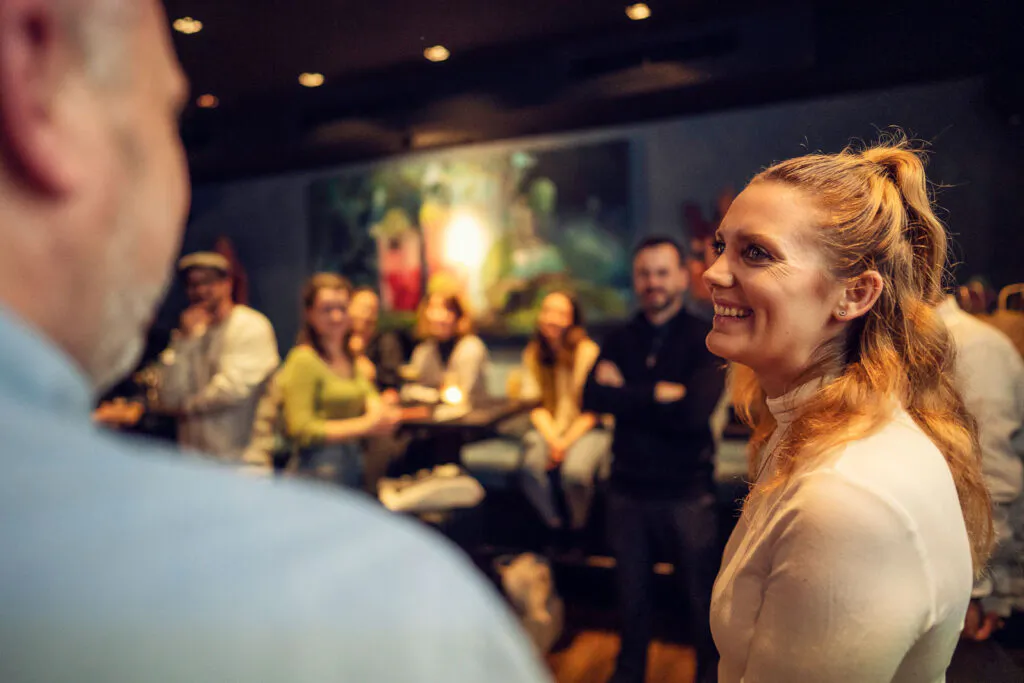Eine lächelnde Frau spricht mit einem Mann, dahinter stehen Gäste in einer warm beleuchteten Bar.