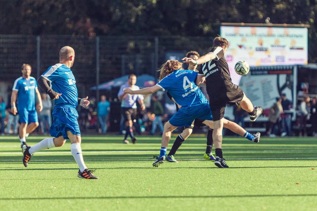 Fußballspieler in blauen und schwarzen Trikots kämpfen bei Sonnenlicht auf einem Kunstrasenplatz um den Ball.