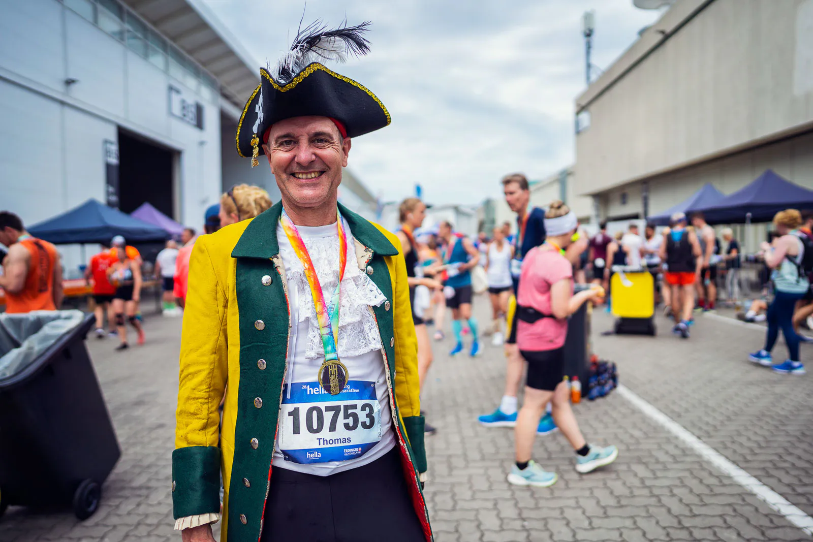 Lächelnder Läufer in Piratenkostüm mit Dreispitz, Startnummer und Medaille zwischen Hallen und anderen Teilnehmenden.
