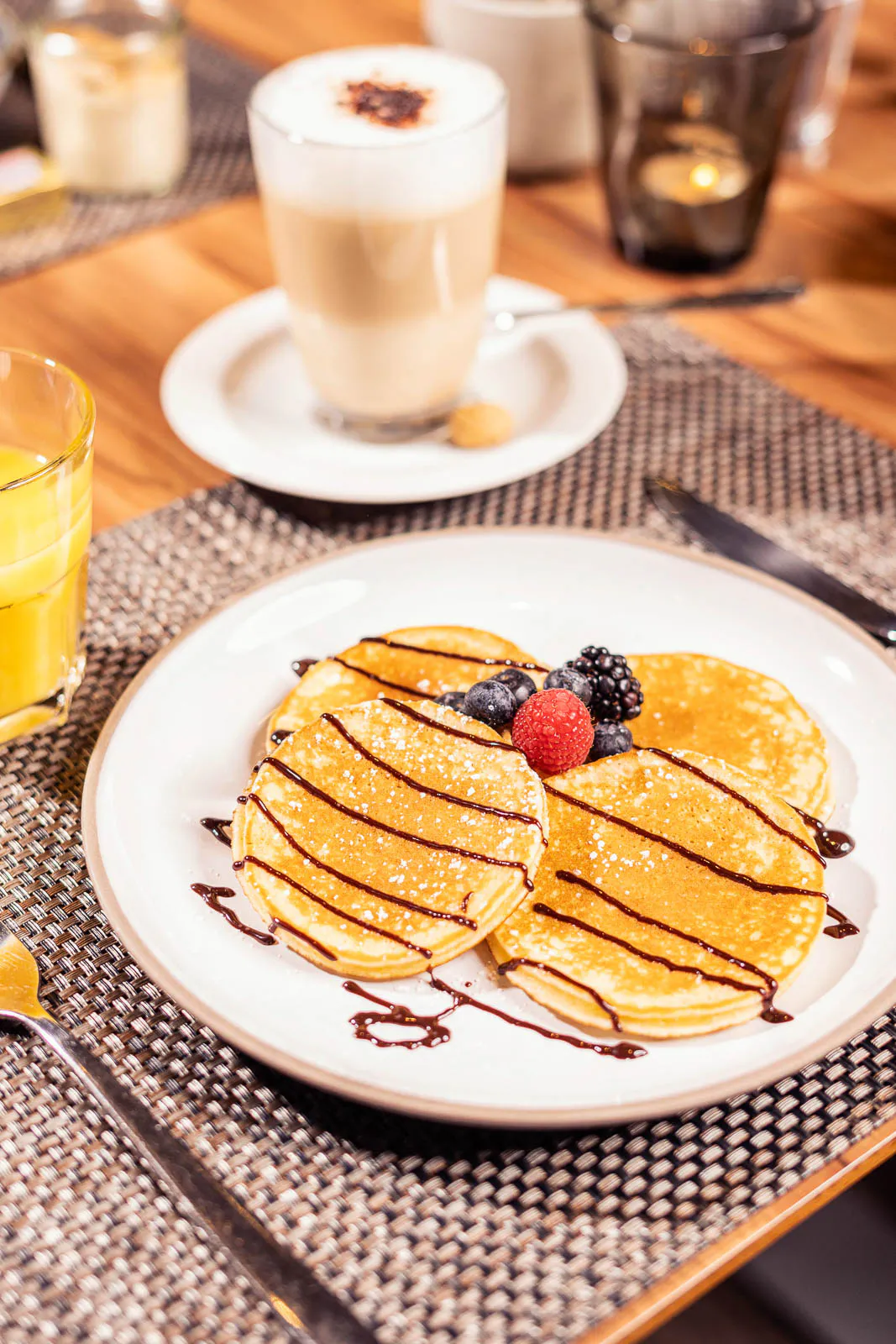 Pancakes mit Schokosauce und Beeren auf weißem Teller, daneben Orangensaft und Latte macchiato auf einem Holztisch.