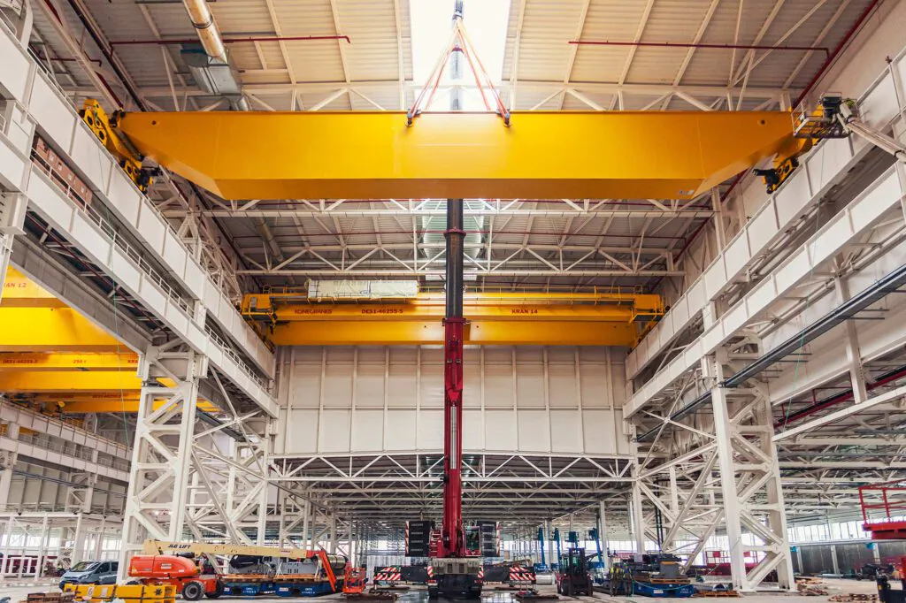 Ein Mobilkran hebt einen gelben Brückenkran in einer hohen, durch Oberlichter beleuchteten Industriehalle.