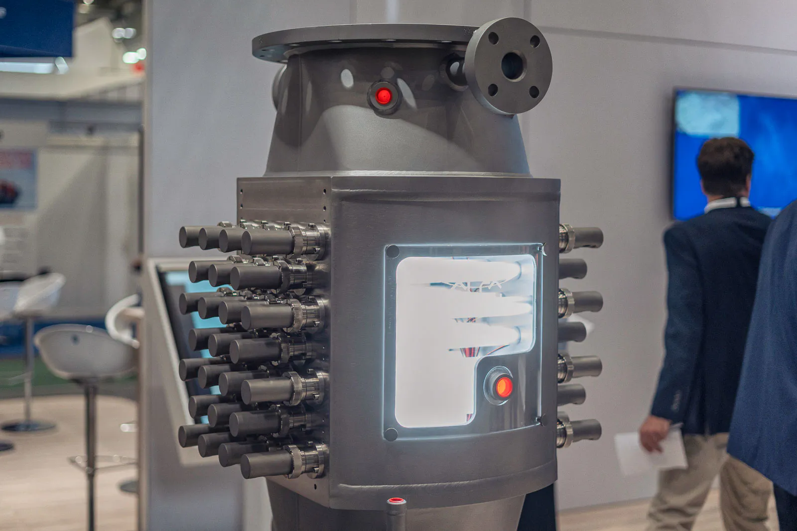 Graues Industriemodell mit Rohranschlüssen und beleuchtetem Sichtfenster am Alfa Laval-Messestand, Besucher im Hintergrund.