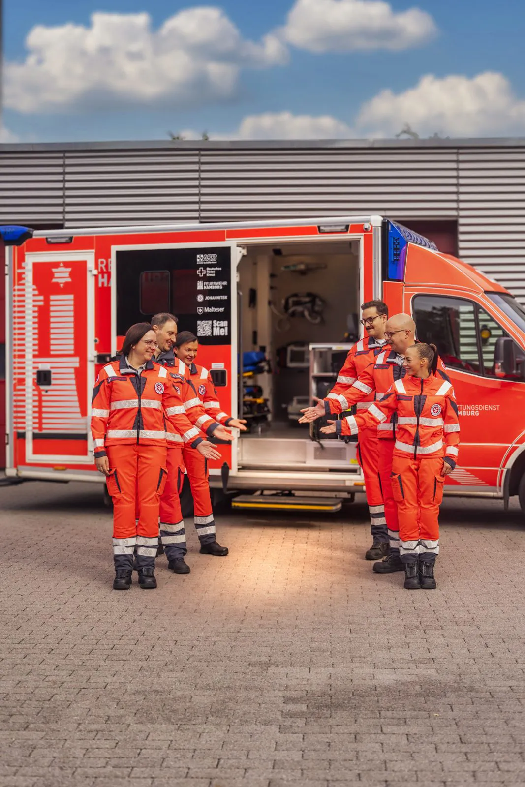 Sechs Johanniter-Rettungskräfte in roten Uniformen stehen bei Tageslicht vor einem Rettungswagen mit offener Seitentür.