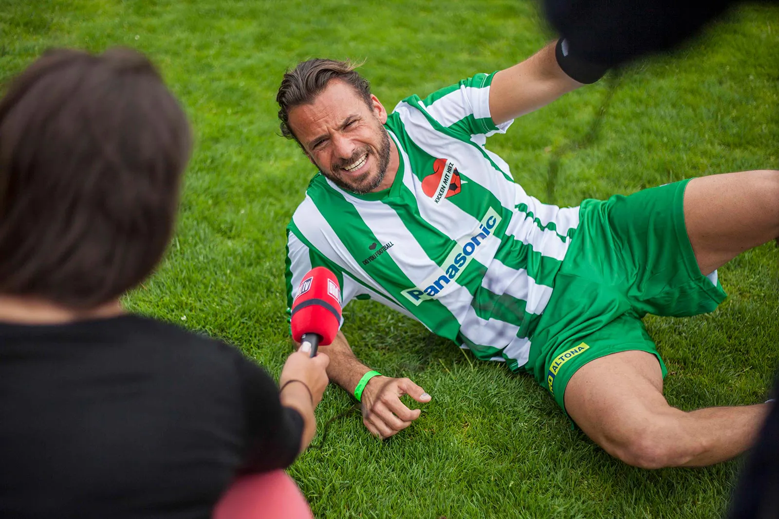Lachender Fußballspieler im grün-weißen Trikot liegt auf dem Rasen und wird mit rotem Mikrofon interviewt.