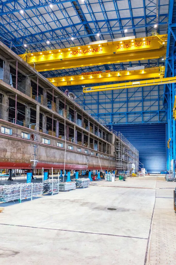 Gelbe Konecranes-Brückenkräne über einem Schiffsrumpf in einer beleuchteten Industriehalle mit blauem Stahltragwerk.