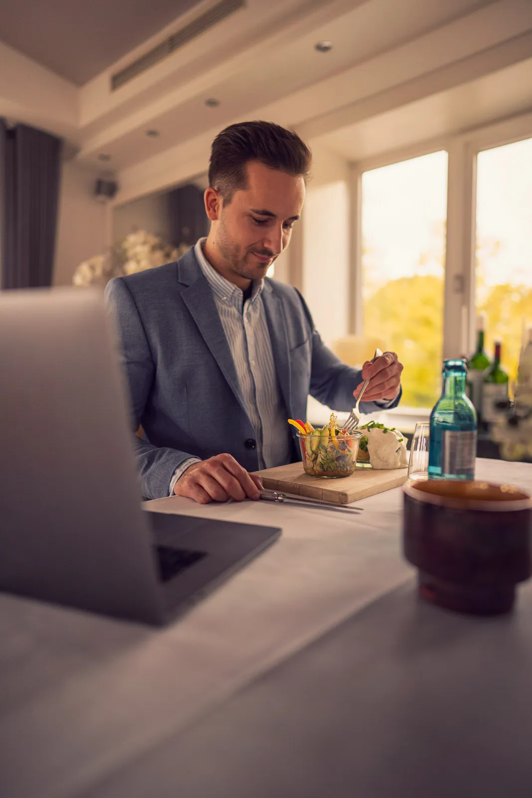 Mann im Sakko isst am Tisch neben einem Laptop; warmes Tageslicht fällt durch die Fenster des Restaurants.