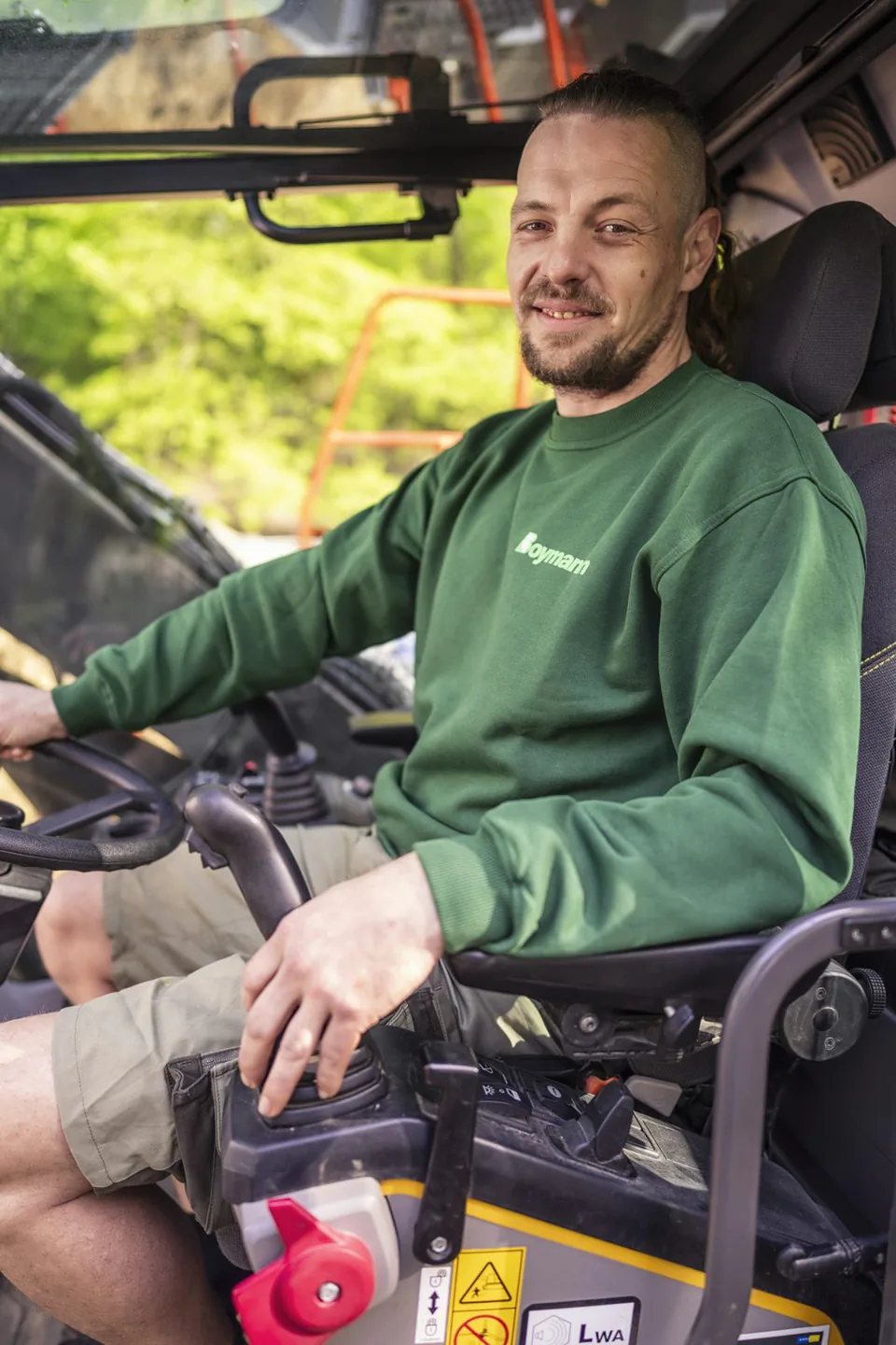Mann im grünen Boymann-Pullover sitzt bei Tageslicht am Steuer einer Arbeitsmaschine, die Hand am Bedienhebel.