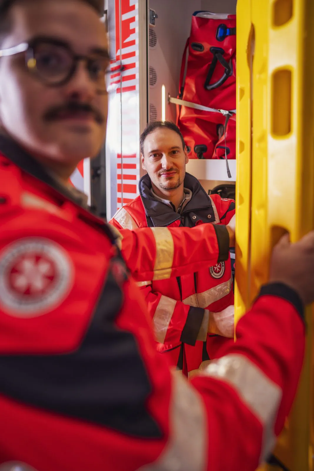 Zwei Johanniter-Rettungskräfte in roten Jacken halten ein gelbes Rettungsbrett im Rettungswagen.