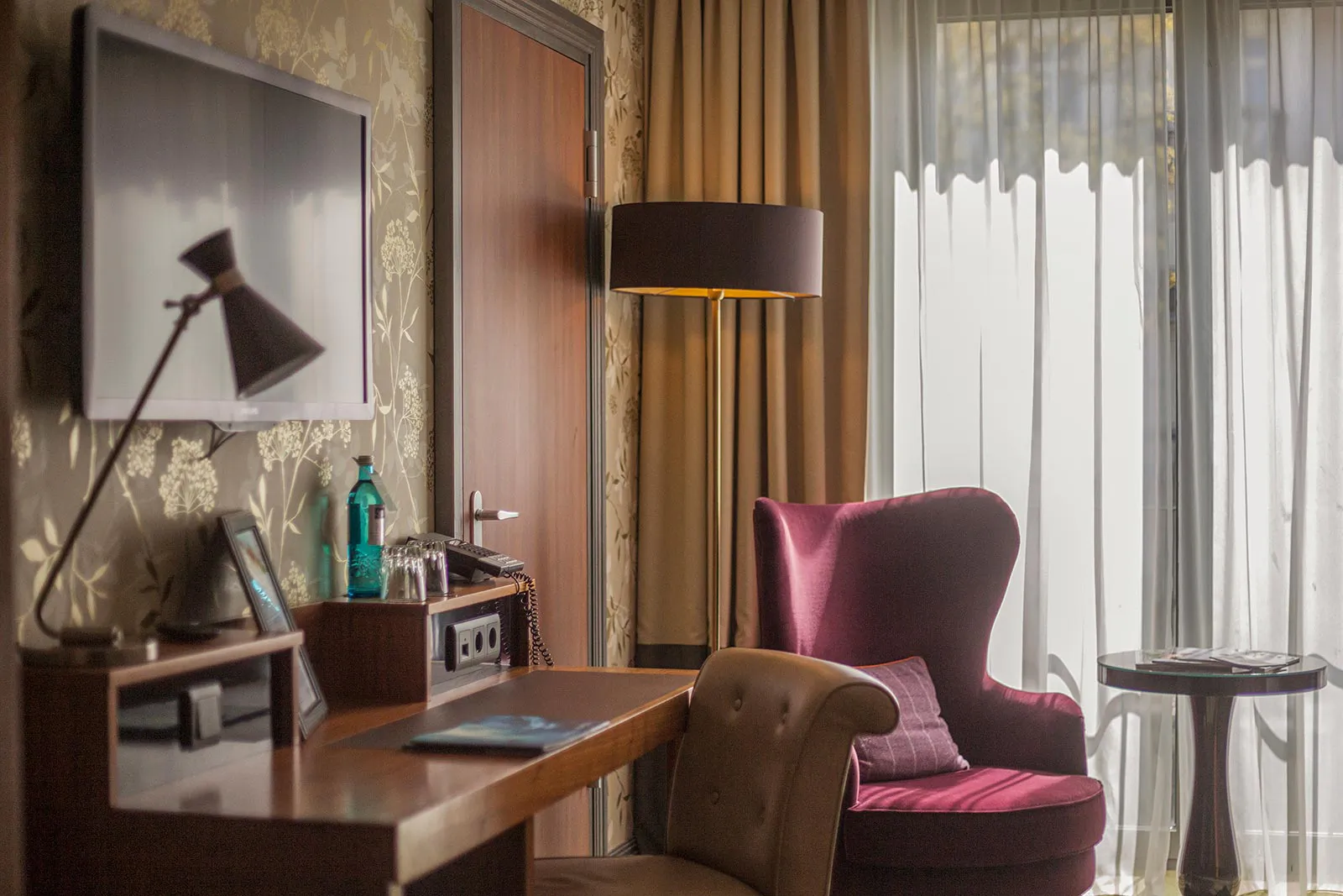 Hotelzimmer mit Schreibtisch, Fernseher und violettem Sessel vor einem großen Fenster im Tageslicht.