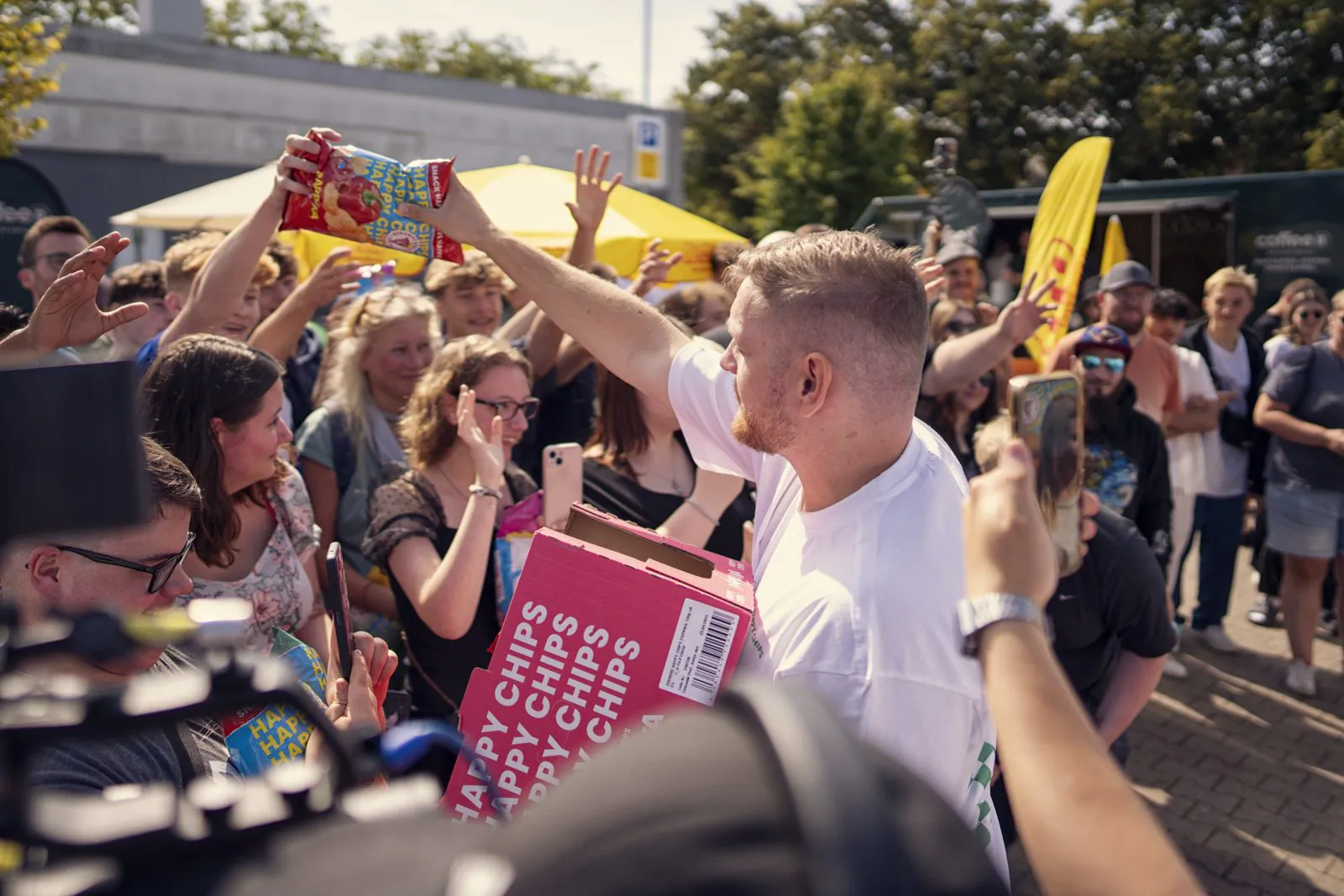 Mann hält eine Happy-Chips-Tüte über eine Menschenmenge beim sonnigen Außenevent, im Arm ein pinker Karton.