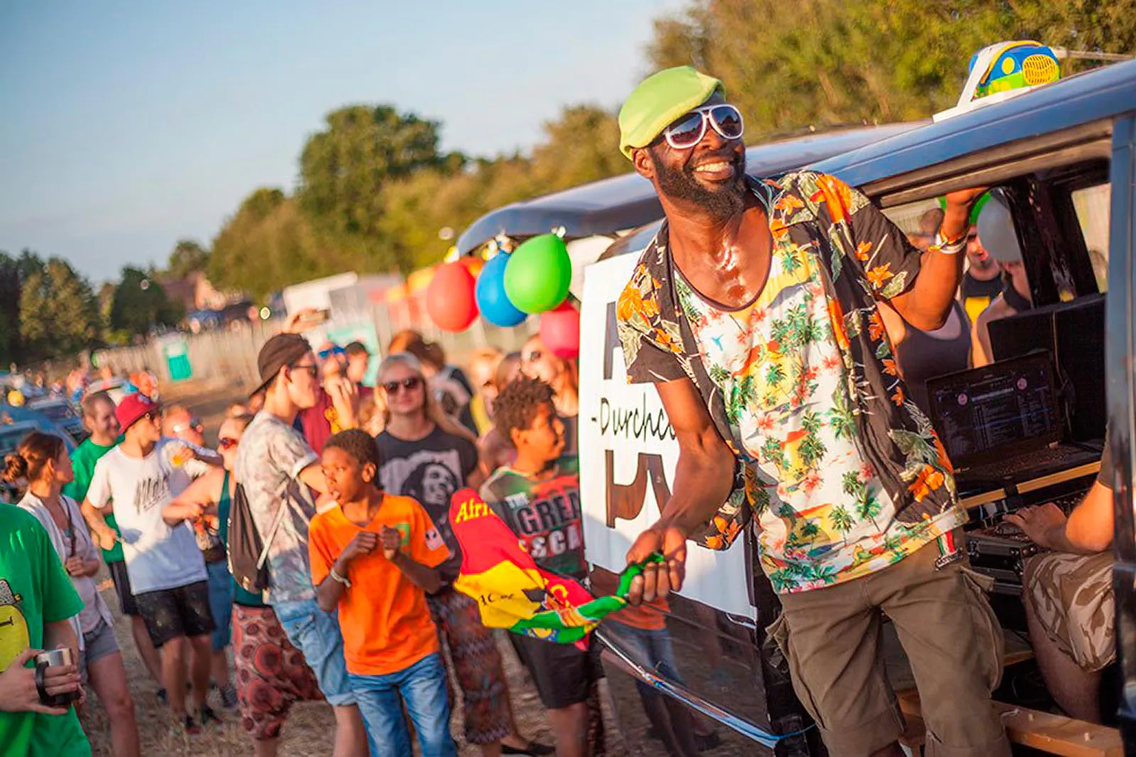 Mann mit grüner Mütze und Sonnenbrille neben einem Van, umringt von Festivalgästen im warmen Abendlicht.