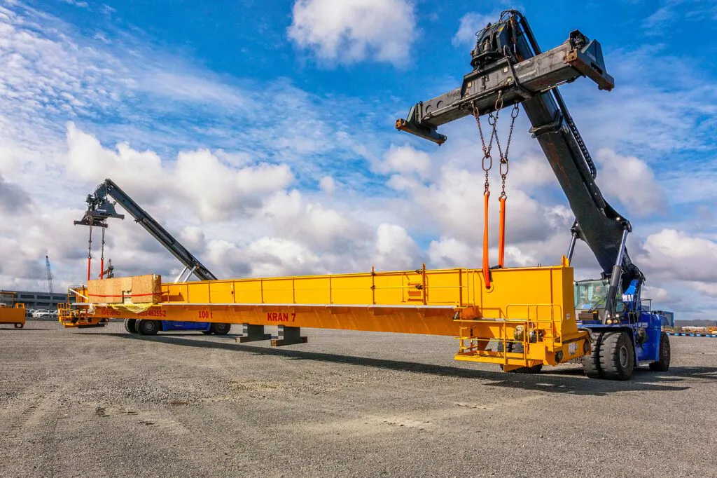 Zwei Reachstacker heben einen langen gelben Kranträger auf einem sonnigen Industrieplatz unter blauem Himmel.