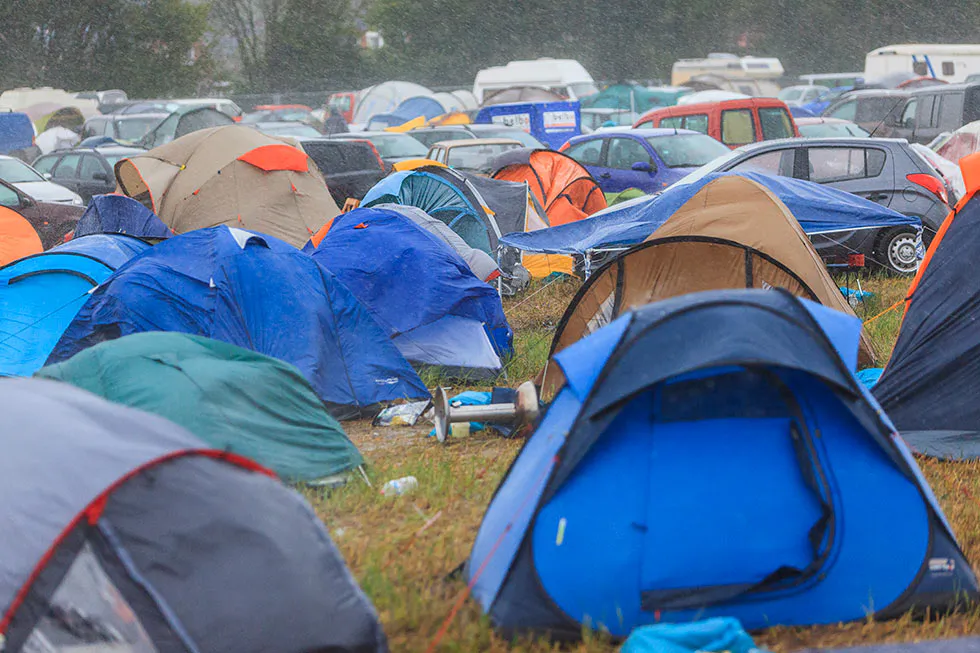 Bunte Zelte dicht an dicht auf einer regennassen Campingwiese, dahinter parkende Autos und Wohnmobile.