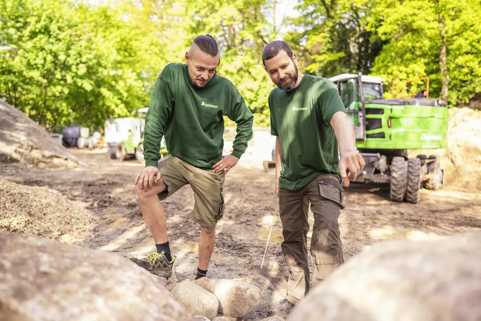 Zwei Männer in grünen Boymann-Shirts begutachten Steine auf einer sonnigen Baustelle, dahinter ein grüner Bagger.