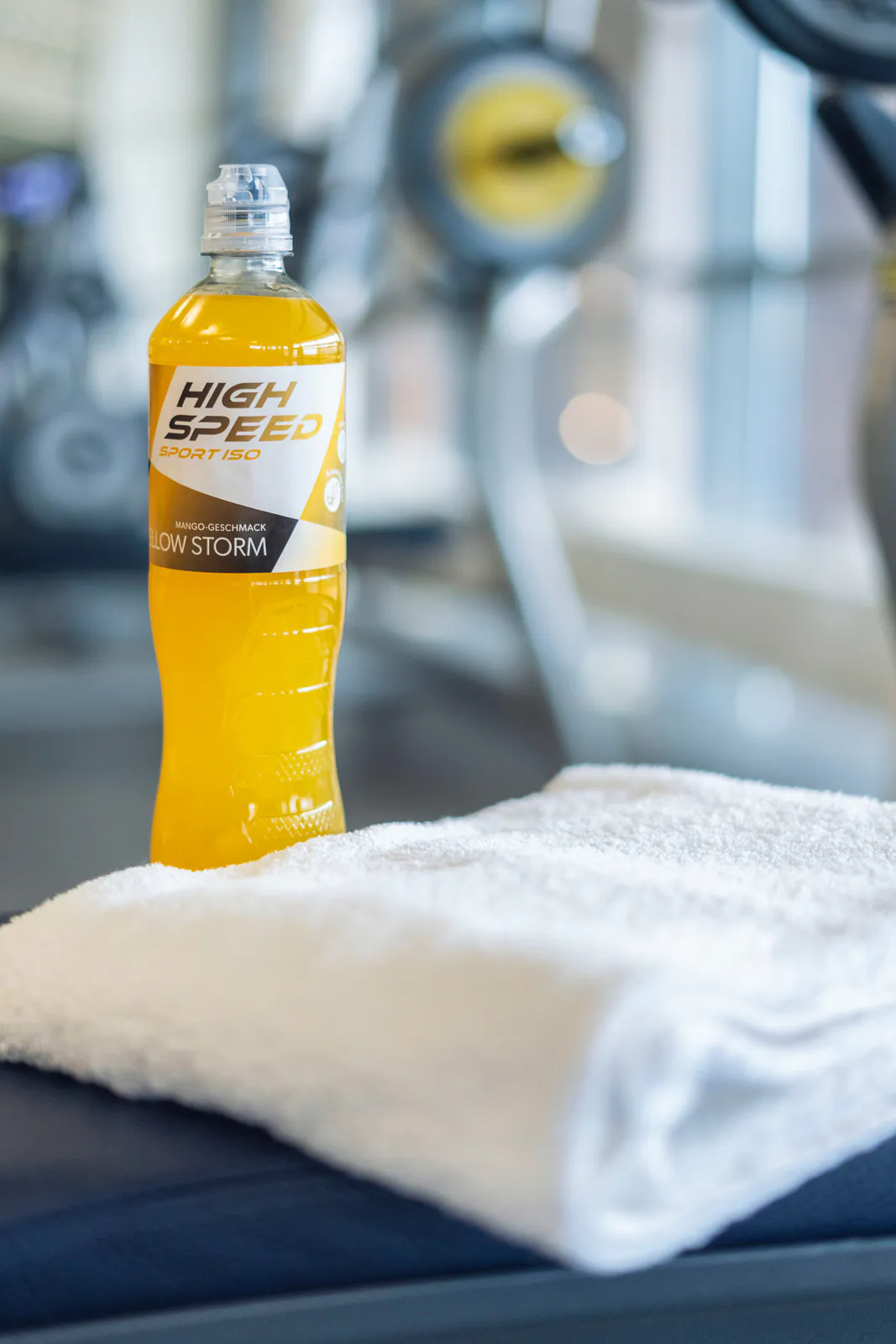 Gelbes Sportgetränk neben einem weißen Handtuch auf einer Trainingsbank im hellen Fitnessstudio.