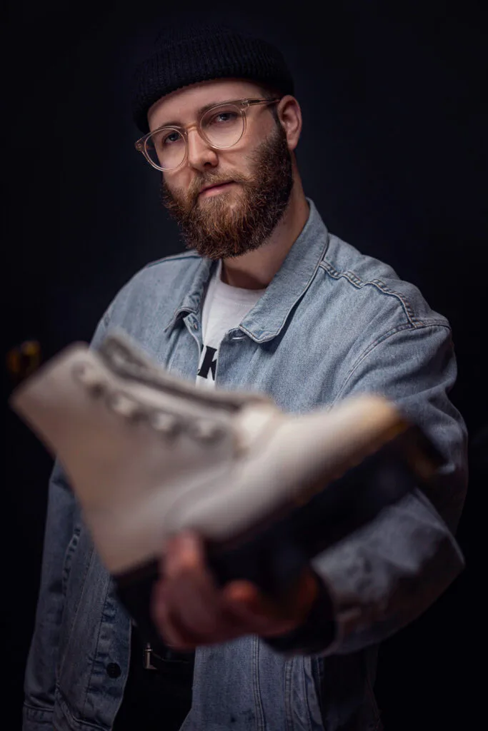Bärtiger Mitarbeiter mit Brille, Mütze und Jeansjacke hält einen hellen Dr. Martens-Stiefel vor dunklem Hintergrund.
