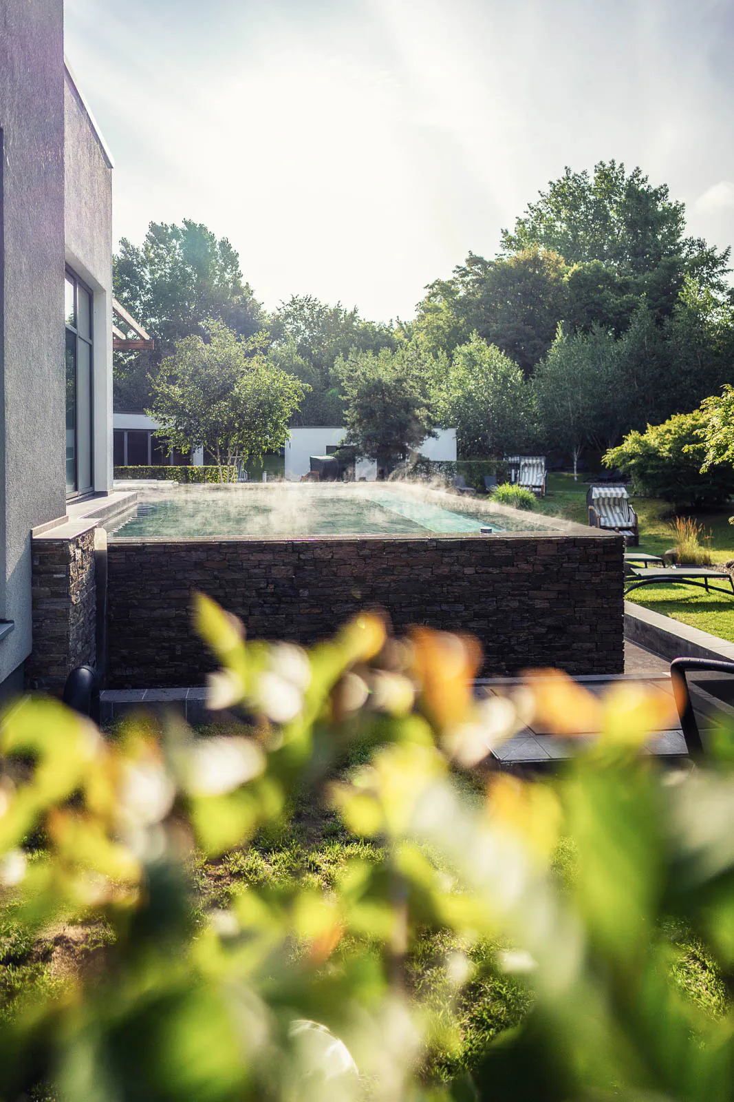 Dampfendes Außenbecken mit Steinverkleidung im sonnigen Saunagarten, umgeben von Bäumen und Liegestühlen.