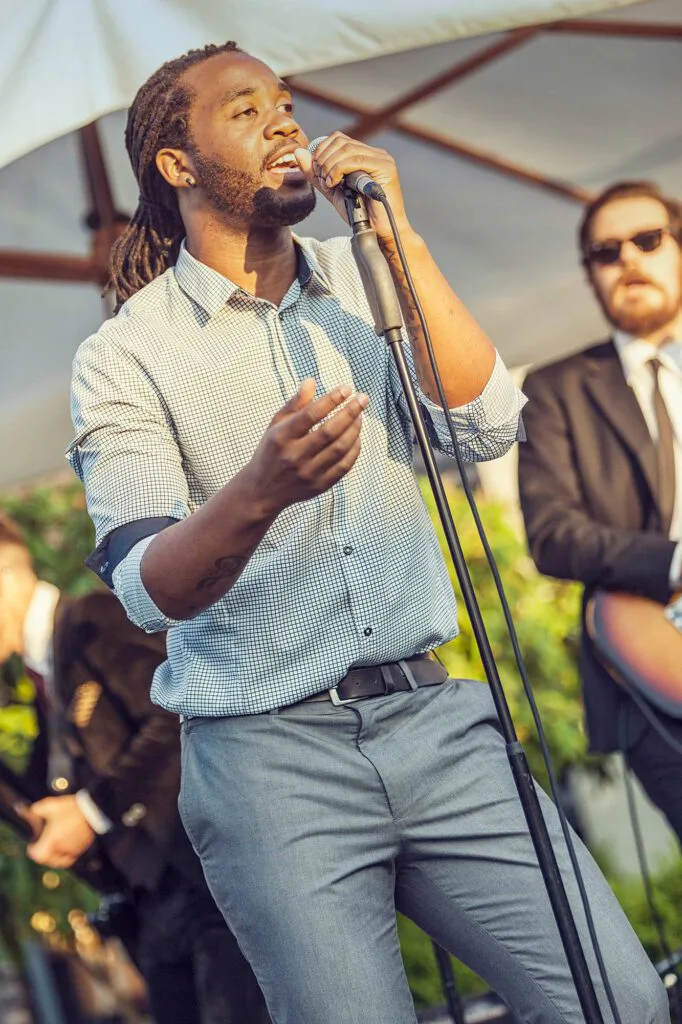 Sänger mit Dreadlocks singt bei Sonnenlicht ins Mikrofon unter einem Zeltdach, rechts ein Gitarrist mit Sonnenbrille.
