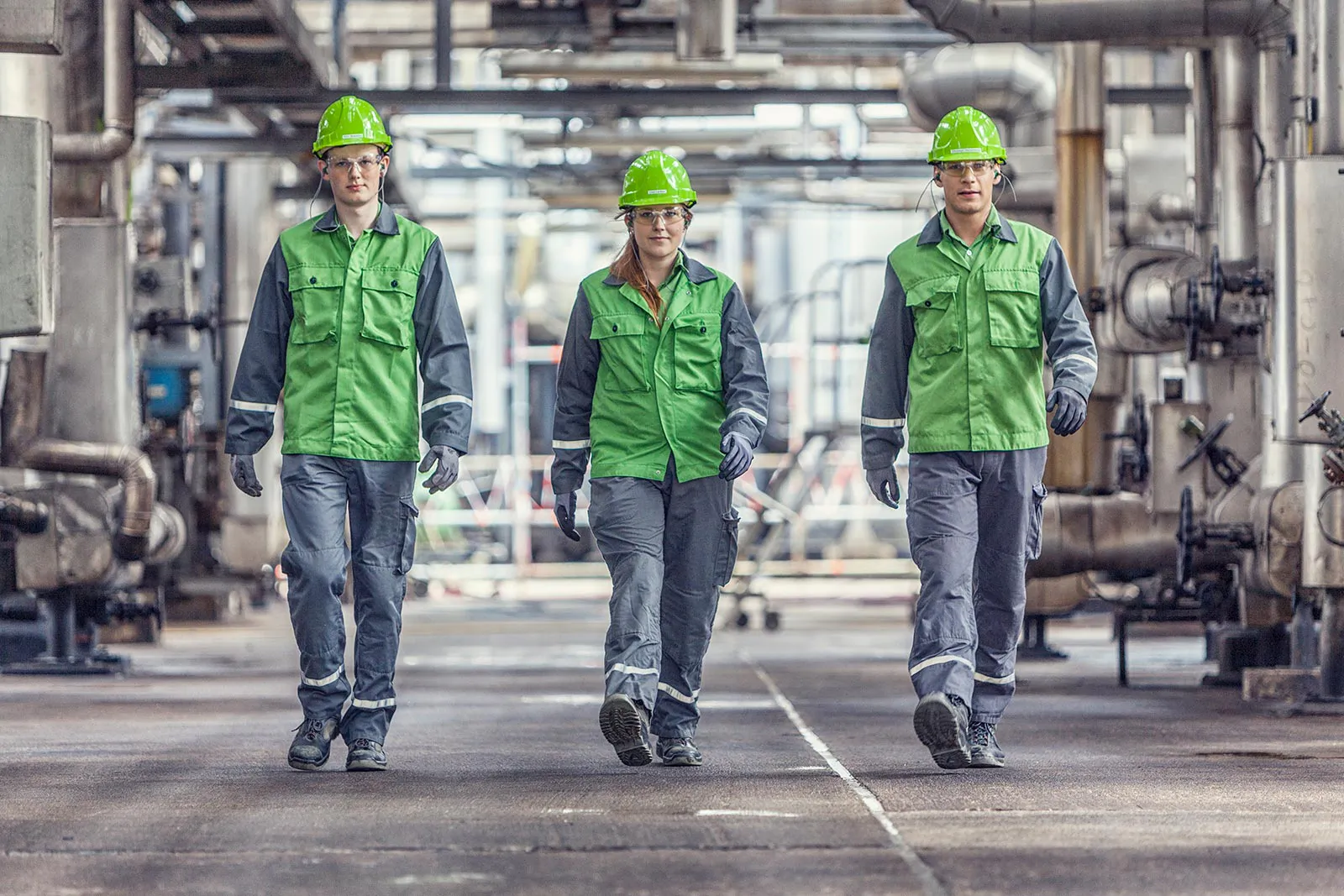 Drei Personen in grüner Schutzkleidung und Helmen gehen zwischen Rohrleitungen durch die Raffinerie Heide.