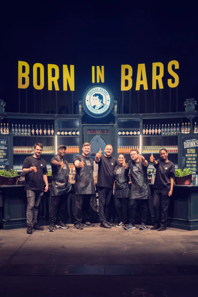 Sieben Personen posieren vor einer beleuchteten Thomas-Henry-Bar mit Flaschenregalen und gelbem „Born in Bars“-Schriftzug.