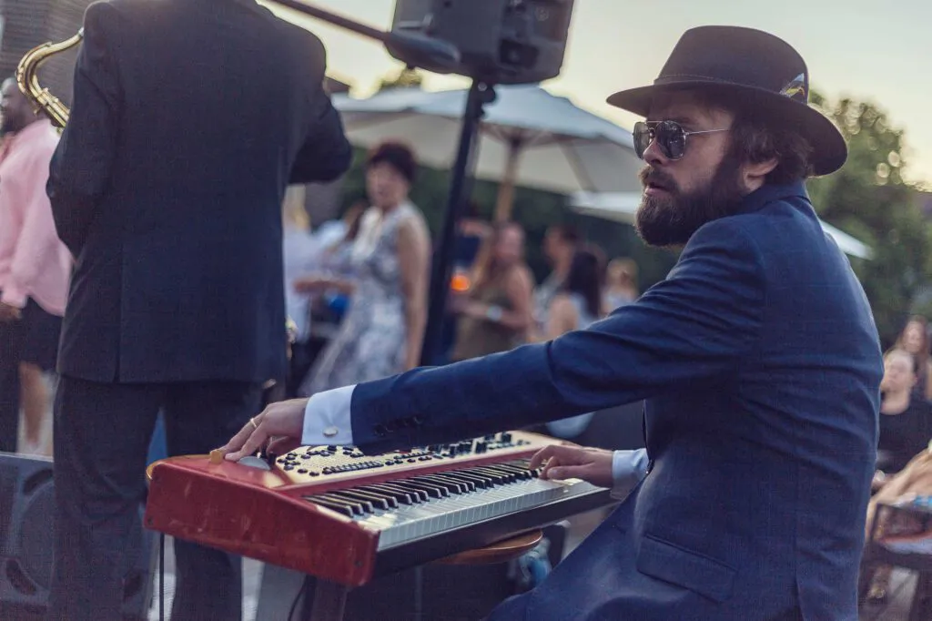 Musiker mit Hut und Sonnenbrille spielt rotes Keyboard im Freien, daneben ein Saxofonist und Publikum im Abendlicht.