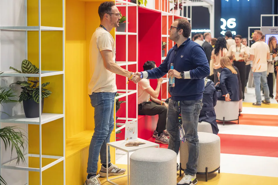 Zwei Männer geben sich am bunt gestalteten Awin-Messestand die Hand, im Hintergrund sitzen und stehen Messebesucher.