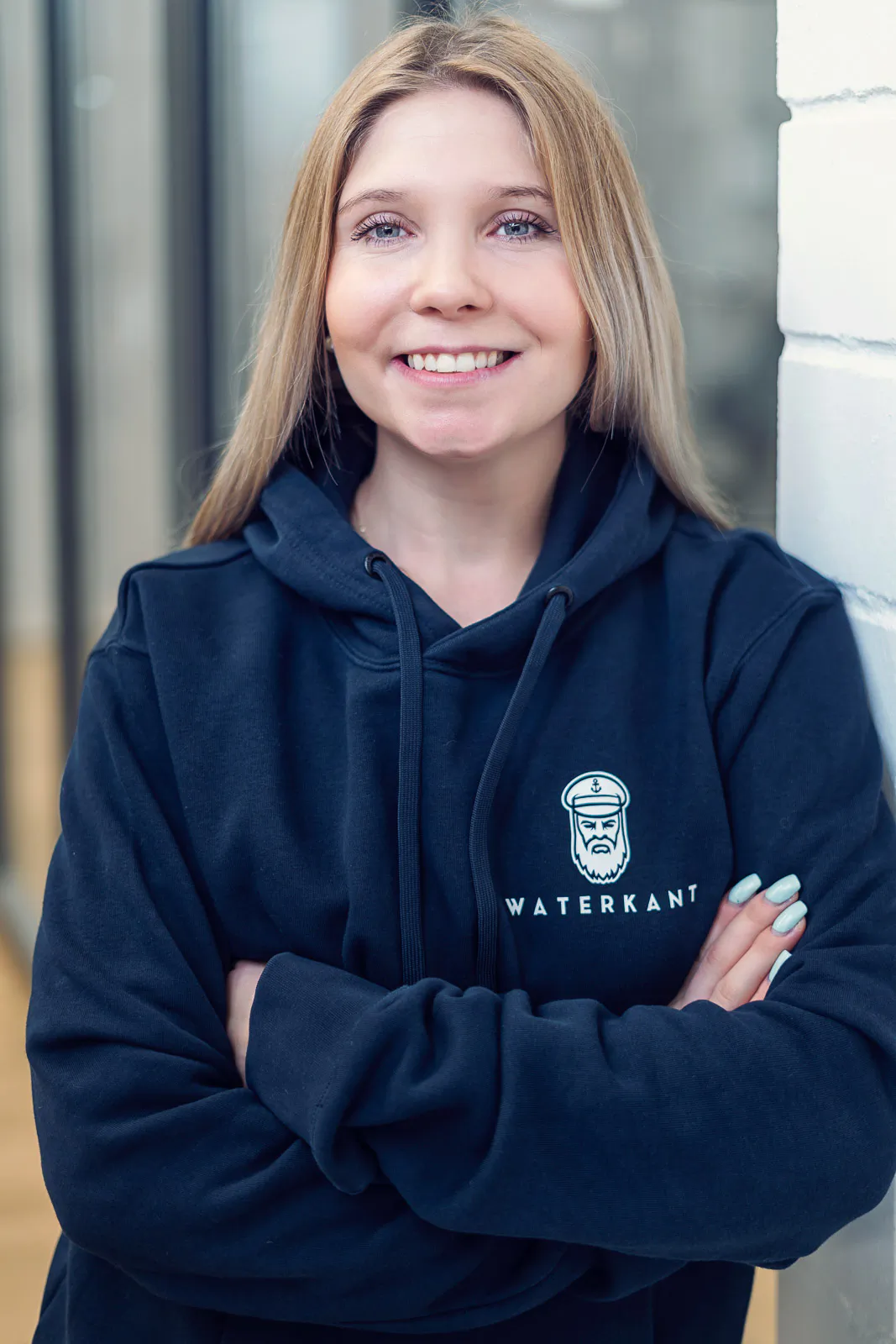 Lächelnde Frau im dunklen Waterkant-Hoodie mit verschränkten Armen an einer weißen Backsteinwand, weich beleuchtet.