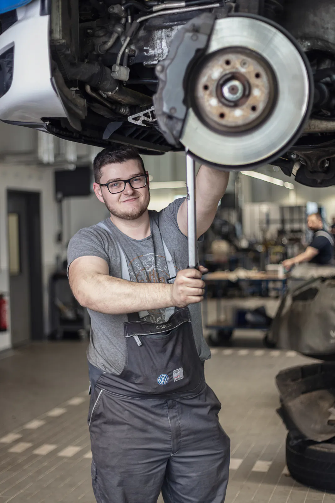 Mechaniker mit Brille arbeitet unter einem angehobenen VW in der Werkstatt, im Vordergrund eine freigelegte Bremsscheibe.