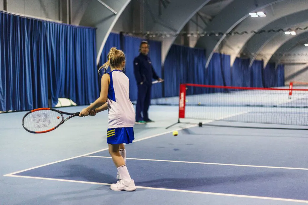 Junge Tennisspielerin mit Schläger am Netz einer Tennishalle mit blauen Vorhängen, im Hintergrund ein Mann.