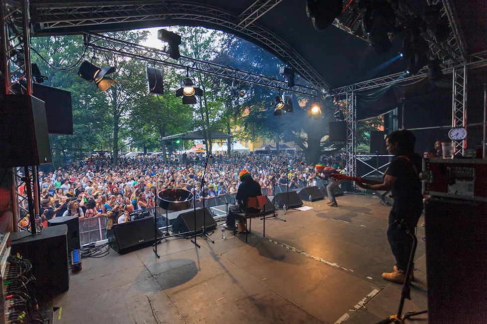 Blick von der überdachten Bühne auf Musiker und dichtes Publikum, mit blauem Scheinwerferlicht und Bäumen im Hintergrund.