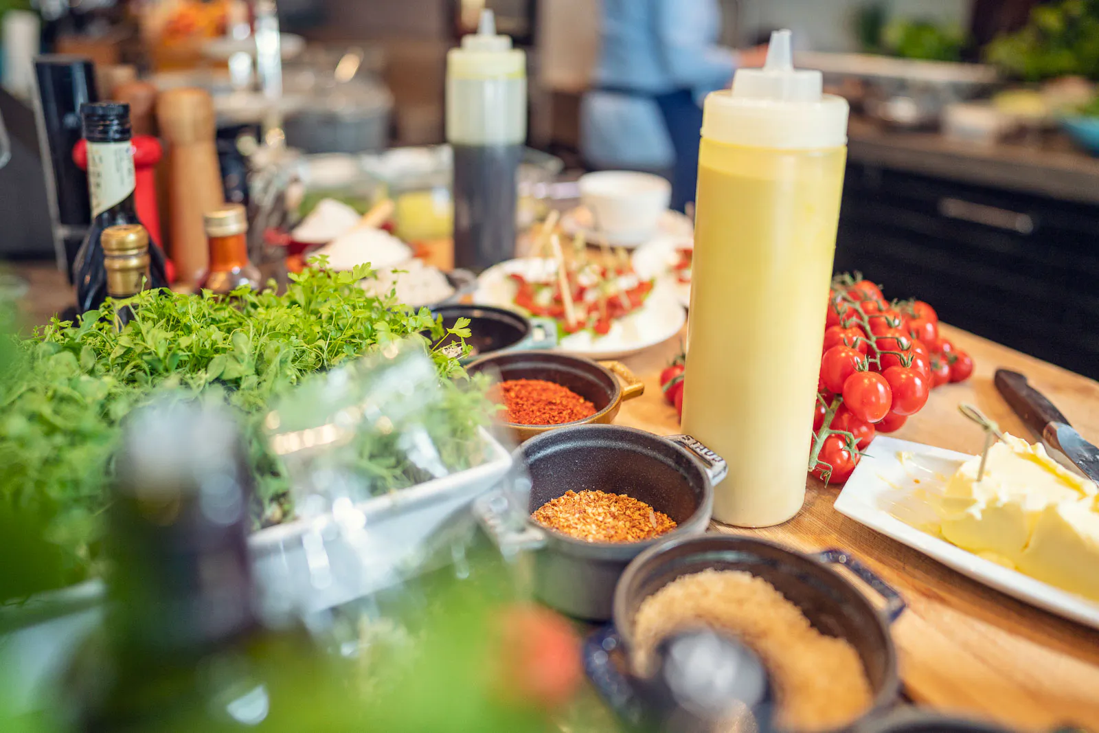 Kräuter, Tomaten, Gewürzschalen, Butter und Dosierflaschen auf einer hell beleuchteten Arbeitsfläche in der Gastroküche.
