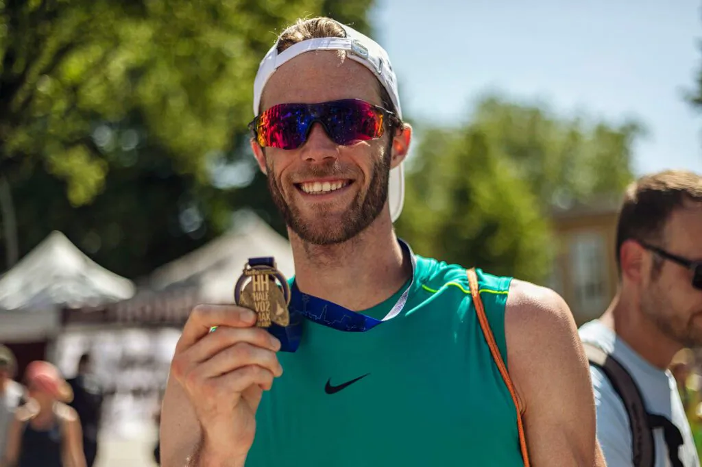 Lächelnder Läufer mit Sonnenbrille und weißer Kappe hält im Sonnenlicht seine Halbmarathon-Medaille hoch.