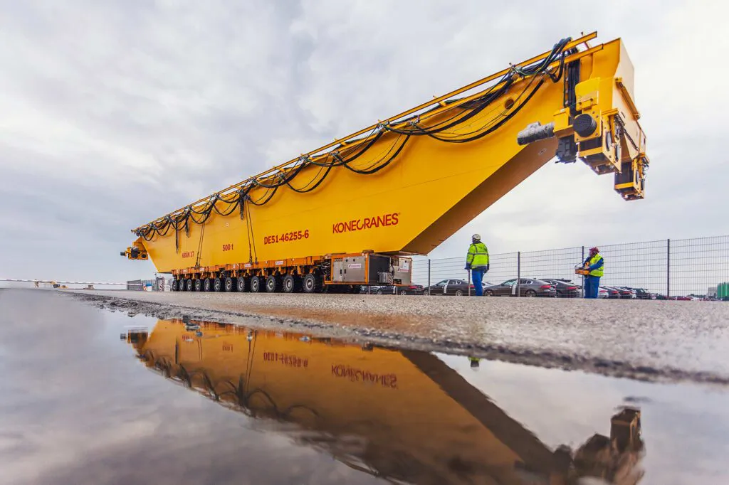 Gelber Konecranes-Kranträger auf Schwertransporter, zwei Arbeiter daneben; Spiegelung in Pfütze unter bewölktem Himmel.
