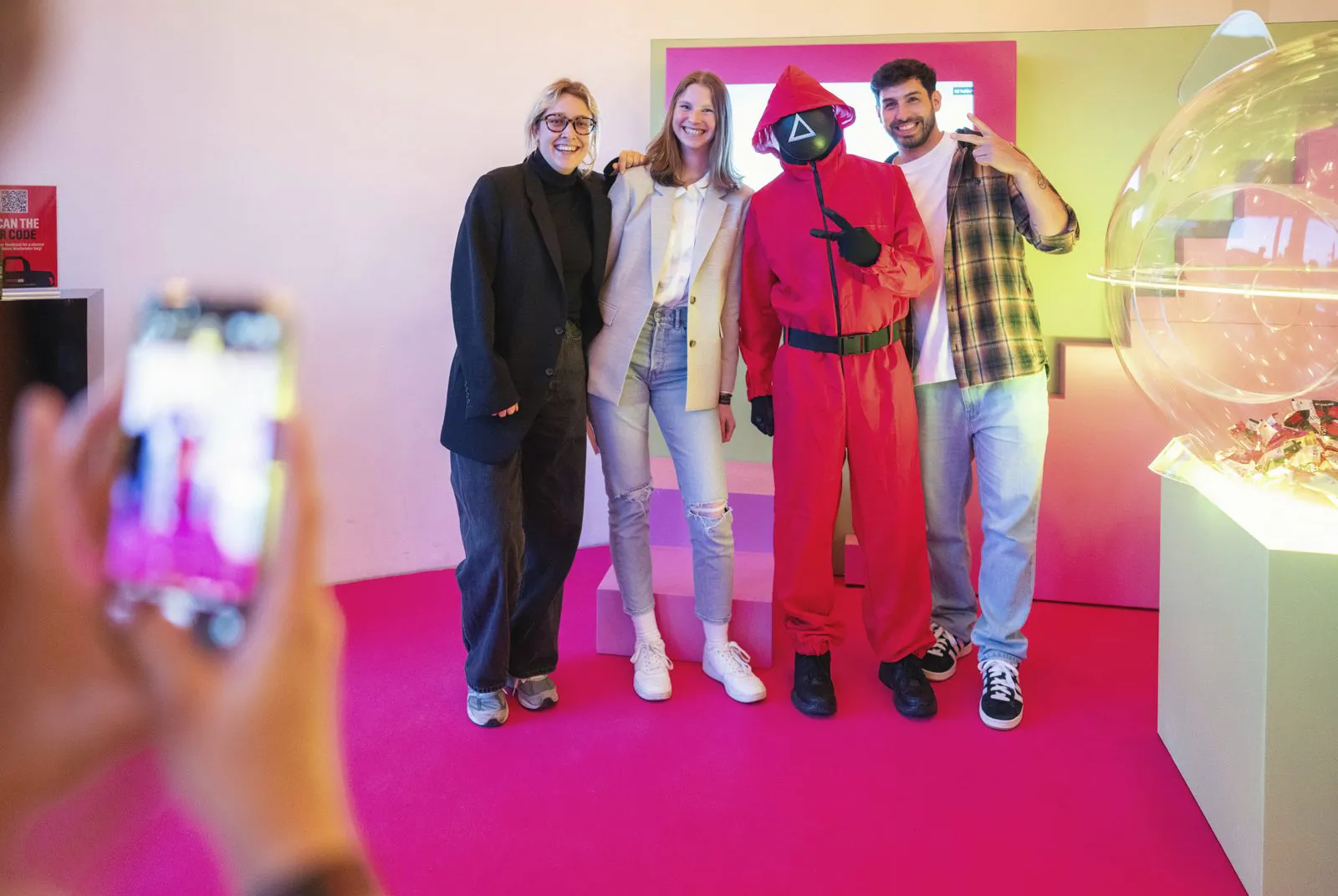 Drei Gäste posieren mit einer rot kostümierten, maskierten Person auf pinkem Teppich beim Netflix-Event.